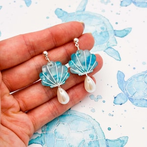 May include: A pair of seashell-shaped earrings. The earrings have a light blue and white design with a silver wire outline and a pearlescent teardrop-shaped bead. The earrings are held in a hand against a background of blue watercolour sea turtles.