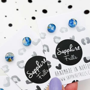 May include: Three pairs of stud earrings with a blue and yellow marbled design, presented on white cards. Each card features a black circle logo with the text "Sapphire Frills". The background is white with black and grey spots.