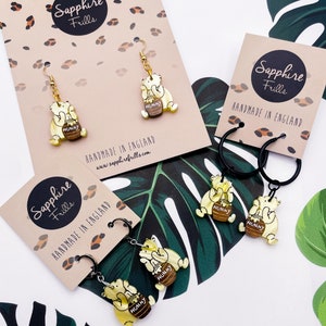 May include: A set of three pairs of Winnie the Pooh inspired earrings. Each pair features a yellow bear holding a honey pot with the word "Hunny" on it. The earrings are made with gold and black metal.
