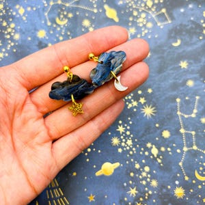 Pu&ograve; includere: Un paio di orecchini a lobo a forma di nuvola marmorizzata blu scuro e nera. Un orecchino presenta un piccolo ciondolo a forma di stella dorata glitterata, mentre l'altro ha un piccolo ciondolo a forma di mezzaluna bianca appeso alla nuvola.