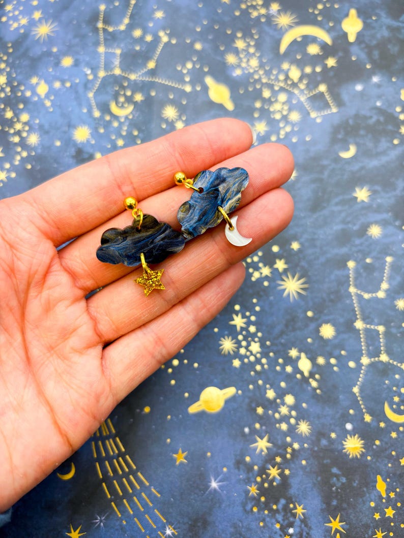 Pu&ograve; includere: Un paio di orecchini a lobo a forma di nuvola marmorizzata blu scuro e nera. Un orecchino presenta un piccolo ciondolo a forma di stella dorata glitterata, mentre l'altro ha un piccolo ciondolo a forma di mezzaluna bianca appeso alla nuvola.