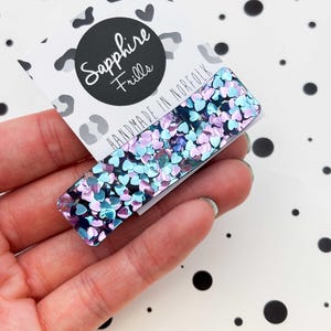 May include: A rectangular hair clip with blue, purple, and pink heart-shaped glitter. The clip is held in front of a card with the text "Sapphire Frills" and "HANDMADE IN NORFOLK" on a white background with black polka dots.