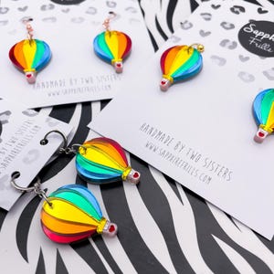 Könnte beinhalten: Ohrringe und Schlüsselanhänger mit bunten Heißluftballons. Die Ballons sind in Regenbogenfarben gehalten, mit roter Basis und goldenen oder silbernen Haken. Der Text "Sapphire Frills" und "Handmade by Two Sisters" ist sichtbar.