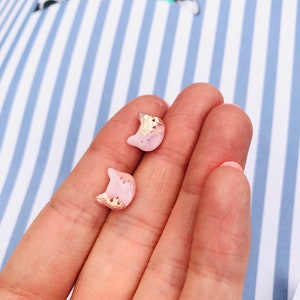Pode incluir: Um par de brincos de lua crescente rosa e ouro. Os brincos são pequenos e delicados, com um design de folha de ouro na frente.