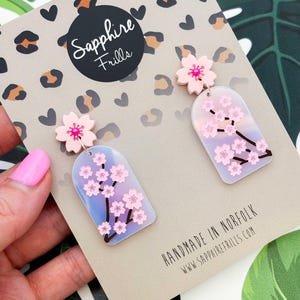 Peut inclure: Boucles d'oreilles fleuries roses et blanches. Elles présentent une fleur rose en haut et un pendentif rectangulaire avec un motif de fleurs de cerisier. Le fond est une carte beige avec le texte "Sapphire Frills" et "Handmade in Norfolk".