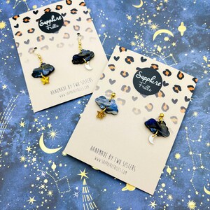 Pu&ograve; includere: Due paia di orecchini pendenti con nuvole marmorizzate blu scuro e nere, stelle glitterate dorate e lune crescenti bianche. Presentati su cartoncini beige con stampa leopardata e il testo Sapphire Frills.