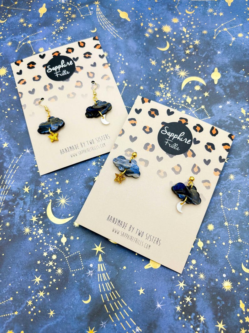Pu&ograve; includere: Due paia di orecchini pendenti con nuvole marmorizzate blu scuro e nere, stelle glitterate dorate e lune crescenti bianche. Presentati su cartoncini beige con stampa leopardata e il testo Sapphire Frills.