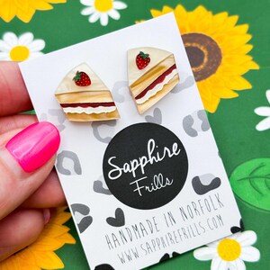 May include: A pair of cake slice earrings with a strawberry on top, displayed on a white card with the text "Sapphire Frills" and "Handmade in Norfolk". The background is a green floral pattern.