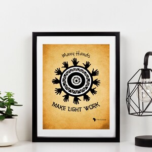 May include: Framed art print with a tan background and black text that reads "Many Hands Make Light Work." The print features a circular design of hands and geometric shapes. A black lamp and a potted plant are also in the image.