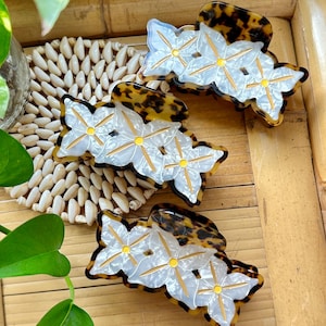 May include: Three flower-shaped hair clips with a tortoiseshell pattern and white and yellow accents. The hair accessories are arranged on a woven mat and bamboo surface, with green foliage in the background.