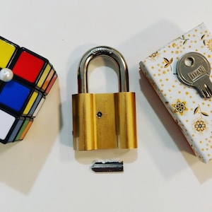 Puede incluir: Un candado dorado con una llave plateada y un pequeño cubo de Rubik. El candado tiene la palabra "TITAN" grabada.