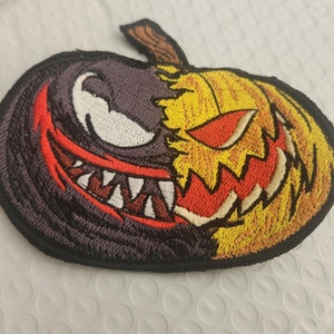 May include: An embroidered patch featuring a split design of a dark purple creature with a wide, toothy grin on the left and a yellow, orange, and red jack-o'-lantern with a jagged, glowing eye and mouth on the right, all outlined in black.