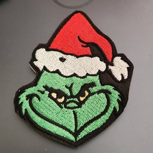 May include: Green and red embroidered patch of the Grinch wearing a Santa hat.
