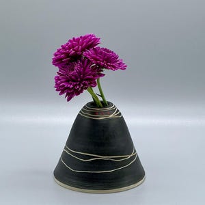 May include: A small black ceramic vase with white lines and a cone shape. The vase is filled with pink flowers.