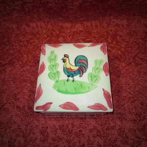 May include: A white ceramic box with a rooster painted on the front. The rooster is yellow, red, and blue with green leaves around it. The box has a red border with white accents.