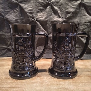 May include: Two black glass beer steins with raised relief designs of a man in a medieval outfit. The steins have a handle on the side and a wide base.