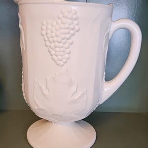 May include: White pitcher with a pedestal base and a large handle. The pitcher features embossed grape and leaf designs. The spout is curved, and the pitcher has a wide opening. The pitcher is made of a solid white material.