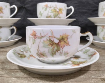 May include: Set of six white porcelain teacups and saucers with a delicate floral pattern of green and orange leaves. The cups and saucers have a gold trim.