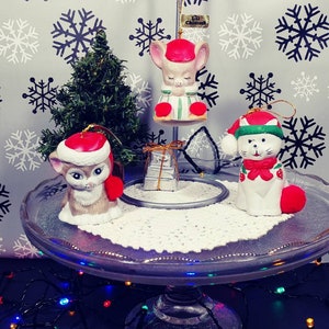 May include: A festive display of Christmas ornaments featuring three ceramic animal figurines: a mouse, a cat, and a kitten, each wearing a Santa hat. The ornaments are arranged on a glass cake stand with a snowflake backdrop.