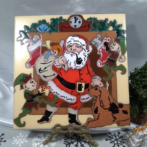 May include: A ceramic Christmas tile featuring Santa Claus in a red suit, holding a sack of toys, with elves and a dog. The background includes a fireplace, clock, and stockings. The tile is displayed on a decorative stand.