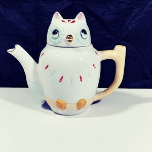 May include: White ceramic teapot shaped like an owl with blue eyes, a gold beak, and red and blue accents. The teapot has a cream-colored handle.