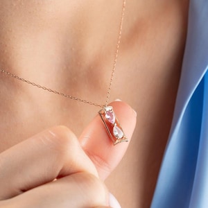 Könnte beinhalten: Eine goldfarbene Halskette mit einem Sanduhr-Anhänger. Der Anhänger hat einen rosa herzförmigen Edelstein oben und einen klaren birnenförmigen Edelstein unten. Die Halskette wird vor einem hellblauen Hintergrund gezeigt.
