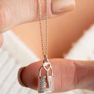Puede incluir: Un collar de oro rosa con un colgante de candado incrustado con piedras de circonita cúbica transparentes. El candado tiene una cerradura en forma de corazón y una llave con una parte superior en forma de corazón.