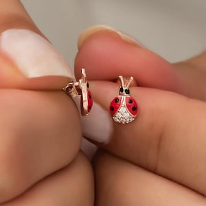 Könnte beinhalten: Ein Paar rote Marienkäfer-Ohrringe mit schwarzen Punkten und einem einzelnen klaren Strassstein auf jedem. Die Ohrringe sind aus goldfarbenem Metall.