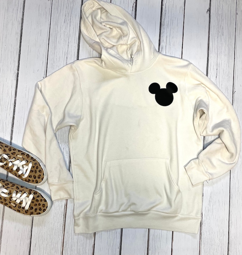 May include: A cream-colored hoodie with a black Mickey Mouse silhouette on the front. The hoodie has a drawstring hood and a kangaroo pocket.