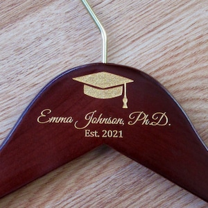 Puede incluir: Una percha de madera marr&oacute;n oscuro con un dise&ntilde;o de gorro de graduaci&oacute;n dorado y el texto "Emma Johnson, Ph.D. Est. 2021" grabado en la parte delantera.