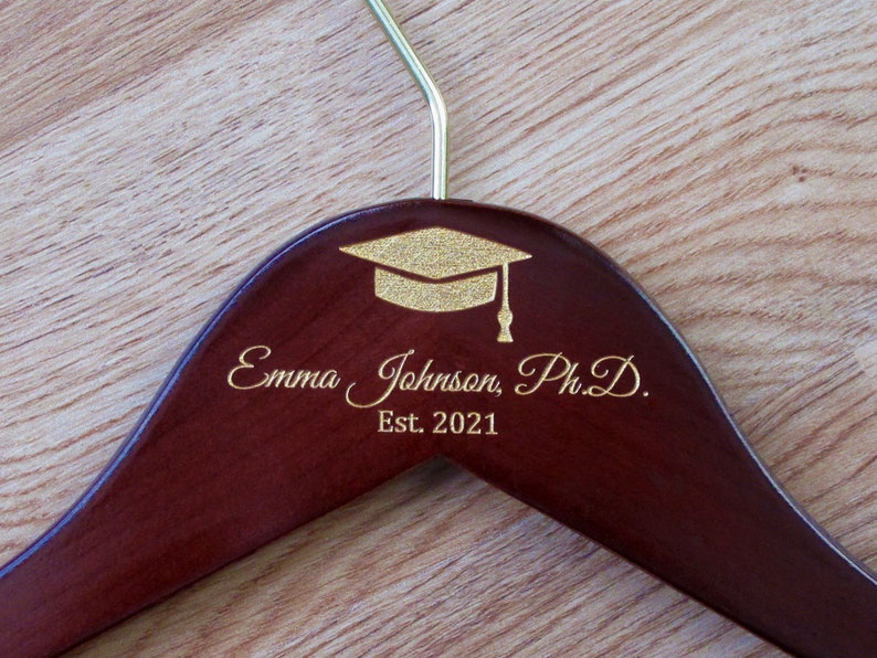 Puede incluir: Una percha de madera marr&oacute;n oscuro con un dise&ntilde;o de gorro de graduaci&oacute;n dorado y el texto "Emma Johnson, Ph.D. Est. 2021" grabado en la parte delantera.