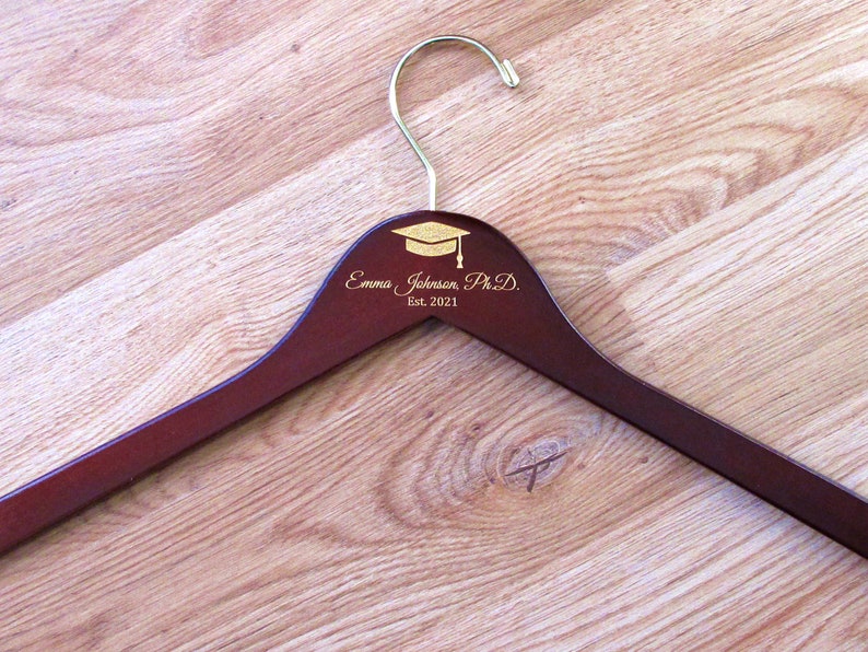 Puede incluir: Una percha de madera marr&oacute;n con un gancho dorado y un dise&ntilde;o de gorro de graduaci&oacute;n dorado. La percha est&aacute; personalizada con el texto "Emma Johanson Ph.D. Est. 2021".
