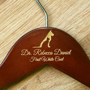 May include: A brown wooden hanger with a gold dog silhouette and the text "Dr. Rebecca Daniel First White Coat" engraved on it.
