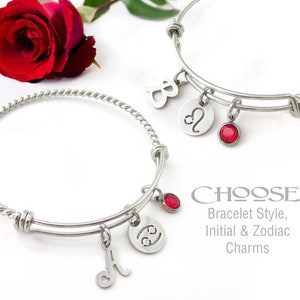 May include: Two silver bangle bracelets with charms. One bracelet has a twisted design and the other is a simple bangle. Both bracelets have a red gemstone charm, a zodiac sign charm, and an initial charm. The text on the image reads "CHOOSE: Bracelet Style, Initial & Zodiac Charms".