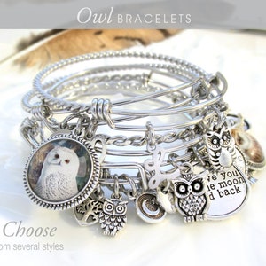 May include: Set of silver bangle bracelets with owl charms. The charms include a white owl, a silver owl, a silver heart with a tree, a silver crescent moon, and a charm with the text "Love you to the moon and back".