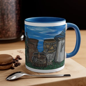 May include: A white ceramic mug with a blue rim and handle. The mug features a hand-painted illustration of four barrels with the names "Legolas", "Gimli", "Boromir", and "Faramir" written on them. The barrels are set against a blue sky background with white clouds.