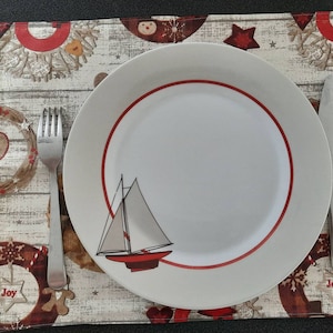 Peut inclure: Une assiette blanche avec un bord rouge et un motif de voilier. L'assiette est posée sur un set de table à motif bois et décorations de Noël rouges et blanches. Une fourchette et un couteau en argent sont placés à côté de l'assiette.