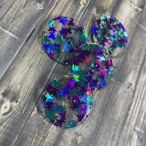 May include: Four round, clear resin buttons with a holographic finish. The buttons are decorated with iridescent purple, green, and blue leaves.