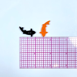 May include: Two fish-shaped buttons, one black and one orange, are shown on a white surface with a ruler for scale. The buttons are approximately 2.5 cm in length.