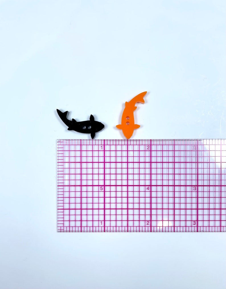 May include: Two fish-shaped buttons, one black and one orange, are shown on a white surface with a ruler for scale. The buttons are approximately 2.5 cm in length.