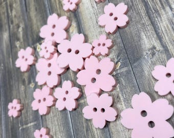 May include: A collection of pink plastic buttons shaped like cherry blossoms. The buttons have two holes each and are scattered on a wooden surface.