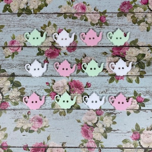 May include: A collection of 10 teapot-shaped buttons in pastel pink, green, and white. The buttons are arranged in three rows on a floral patterned background.