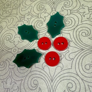 May include: A collection of decorative buttons in a festive arrangement. The image features three green holly leaf-shaped buttons and three red, round buttons. The buttons are set against a patterned background, suggesting a holiday or craft theme.