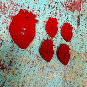 May include: A selection of red heart-shaped buttons. The buttons are of varying sizes, with one large and four smaller. Each button features two sewing holes. They are displayed against a textured, multi-coloured background.