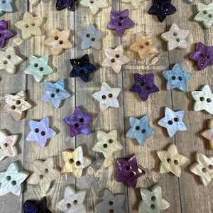 May include: A collection of star-shaped buttons in various colors, including purple, blue, green, and white. The buttons have a pearlescent finish and are arranged on a wooden surface.