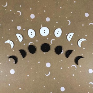 May include: A collection of black and white moon-shaped buttons. The buttons are arranged in a crescent shape on a brown background with white stars.