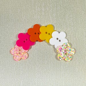 May include: A set of six flower-shaped buttons in a rainbow color scheme. The buttons are made of plastic and have a glossy finish. The buttons are arranged in a semi-circle, with the pink button at the bottom left and the white button at the top right.