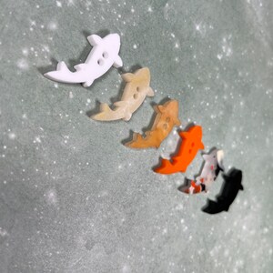 May include: Five colourful fish-shaped buttons with two holes each. The buttons are white, beige, orange, red and black.
