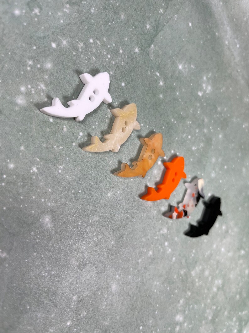 May include: Five colourful fish-shaped buttons with two holes each. The buttons are white, beige, orange, red and black.
