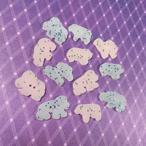 May include: A collection of decorative buttons in the shape of polar bears. The buttons are in shades of pink and light blue, with glitter accents. The buttons have two holes for sewing and are scattered on a purple background.